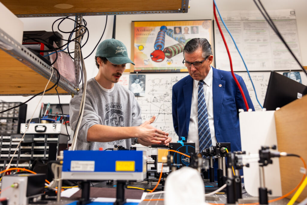 Two individuals examine a complex scientific setup in a laboratory filled with equipment and diagrams.