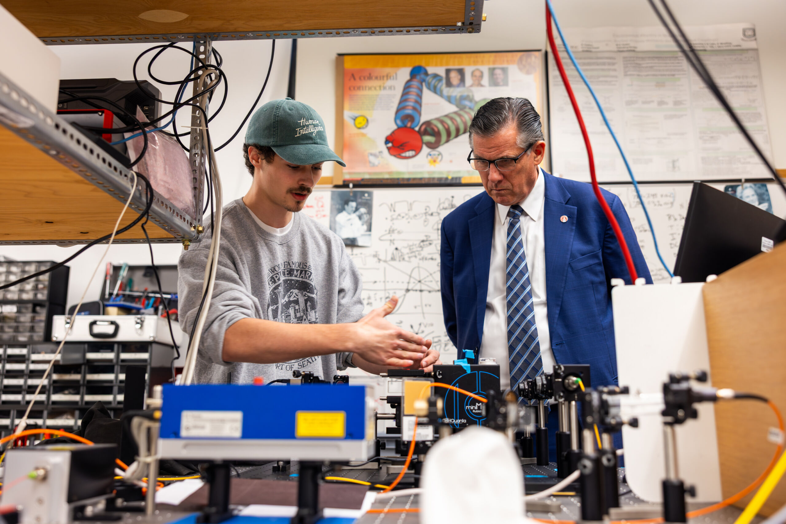 Two individuals examine a complex scientific setup in a laboratory filled with equipment and diagrams.